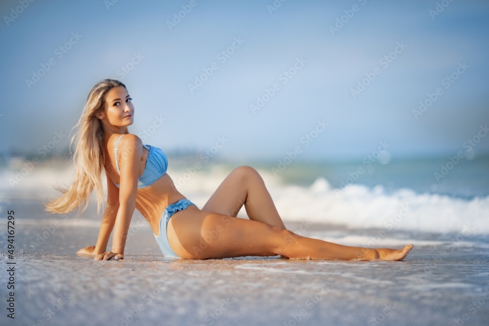 A girl with blond hair in a delicate swimsuit sits by the sea, enjoying the splash of waves A girl with blond hair in a delicate swimsuit sits by the sea, enjoying the splash of waves