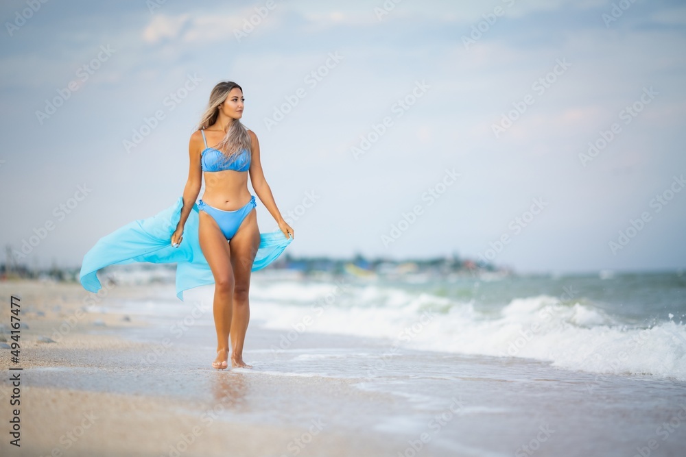 A girl with blond hair in a bluish swimsuit and a bright shawl walks along the beach A girl with blond hair in a bluish swimsuit and a bright shawl walks along the beach