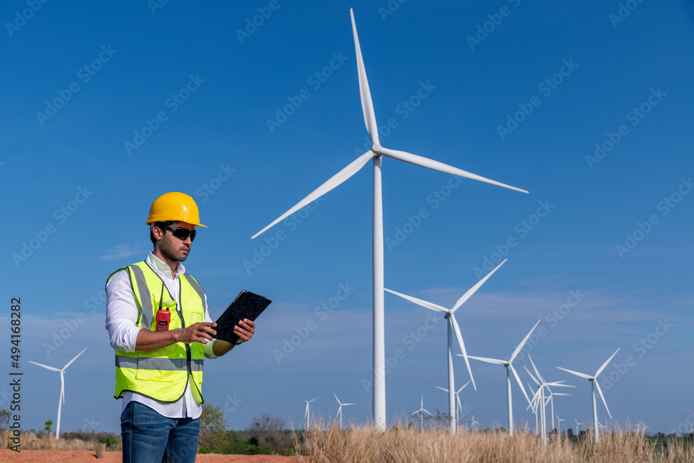 Engineer wearing uniform ,helmet hold document inspection work in wind turbine farms rotation to generate electricity energy. Green ecological power energy generation wind sustainable energy concept. Engineer wearing uniform ,helmet hold document inspection work in wind turbine farms rotation to generate electricity energy. Green ecological power energy generation wind sustainable energy concept.