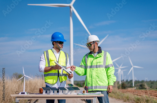 Engineer wearing uniform ,helmet hold document inspection work in wind turbine farms rotation to generate electricity energy. Green ecological power energy generation wind sustainable energy concept. photo