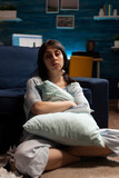 Portrait of depressed woman with mental health problems sitting on floor and hugging pillow in living room. Anxious sad person dealing with chronic disease difficulties and depression at home.