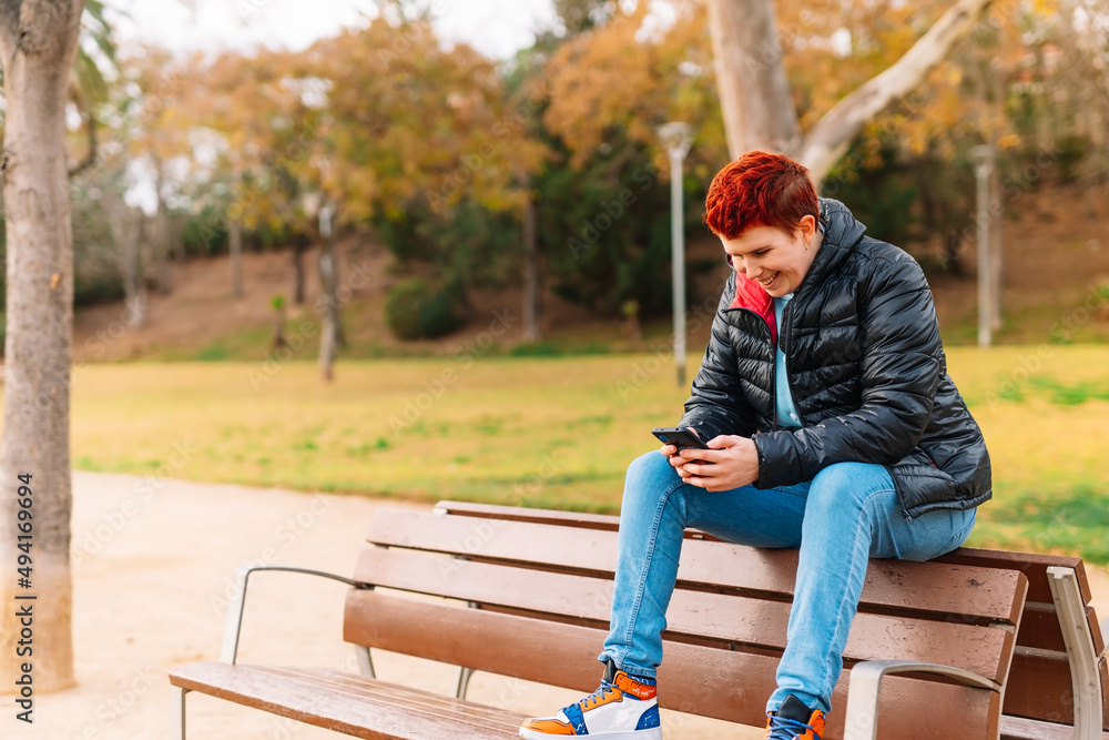 General shot of a modern woman with short red hair, non-binary, looking at her social networks in a public park. General shot of a modern woman with short red hair, non-binary, looking at her social networks in a public park.