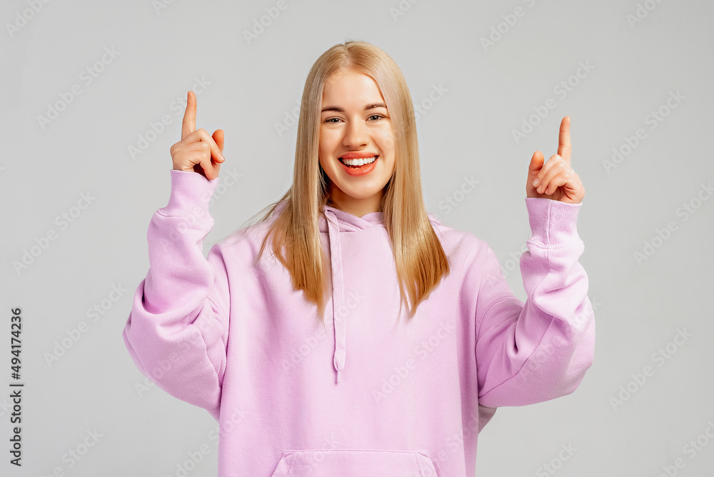 Happy stylish blond girl in trendy hoody pointing fingers up and smiling, inviting to register, showing company logo, standing over gray background, provide information Happy stylish blond girl in trendy hoody pointing fingers up and smiling, inviting to register, showing company logo, standing over gray background, provide information