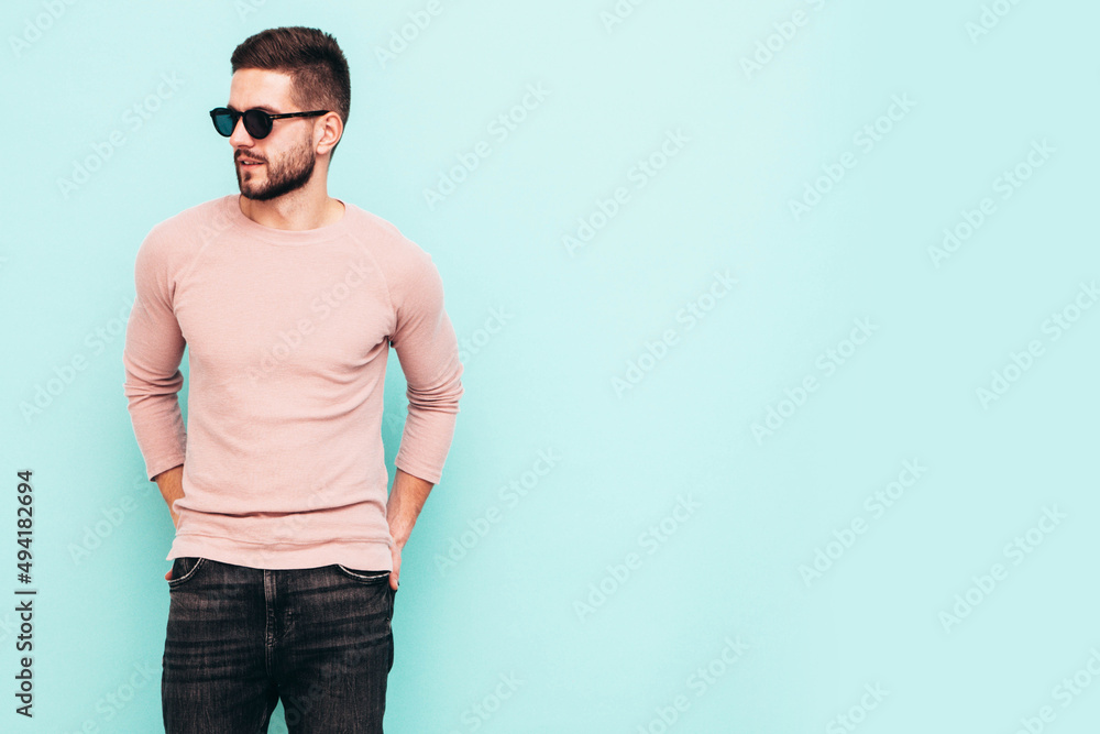 Portrait of handsome confident model. Sexy stylish man dressed in pink sweater and jeans. Fashion hipster male posing near blue wall in studio. In sunglasses. Isolated Portrait of handsome confident model. Sexy stylish man dressed in pink sweater and jeans. Fashion hipster male posing near blue wall in studio. In sunglasses. Isolated
