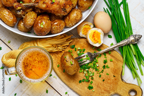 tasty food in rural style, - glass of beer, fried chicken meat and potatoes, garlic and green onions, cheese, boiled eggs and salt, cooked food on a white wooden boards