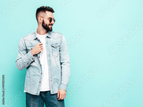 Portrait of handsome confident stylish hipster lambersexual model.Man dressed in jacket and jeans. Fashion male posing near blue wall in studio in sunglasses. Thoughtful. Isolated