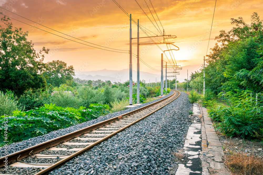 old evening railroad leading to a sunset glow in mountains with green bushes and cjlorful cloudy sku on the background