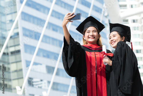 Two happy Asian young beautiful graduate female students with University degree standing and holding diploma taking selfie picture with mobile phone after graduation. Blur background of building