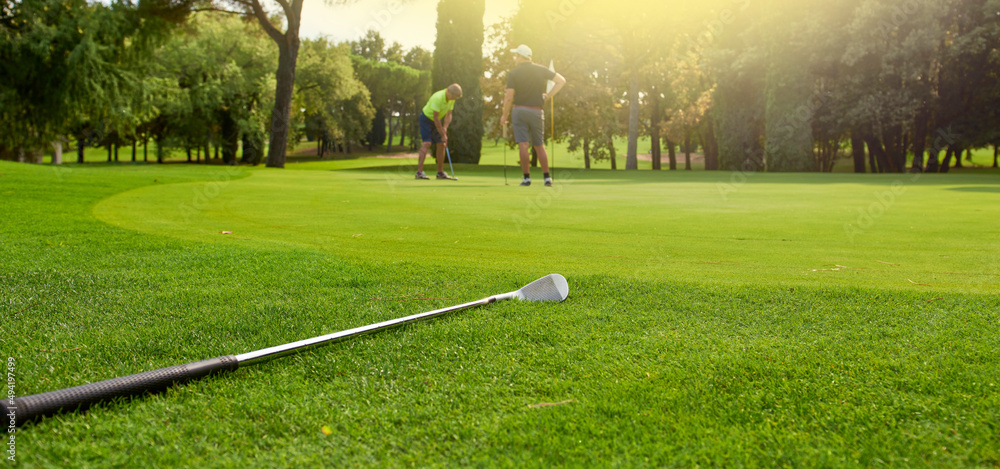 Obraz premium Golfer on the green with a putter in his hands, tries to knock the ball into the hole. A player on the green evaluates the slopes and distance from the hole before aiming the ball towards the flag.