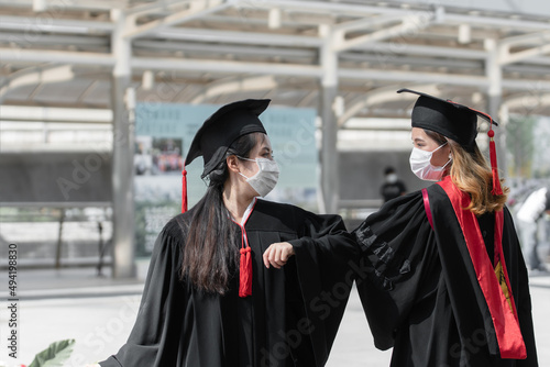 Two Asian young graduate female students wearing face mask standing with elbow greeting at University with black gown, cap with red tassels after graduation. Social distance and new normal concept
