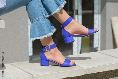 Women's legs in blue denim jeans and sandals in the city street. Trendy elegant casual outfit. Details of everyday summer look.
