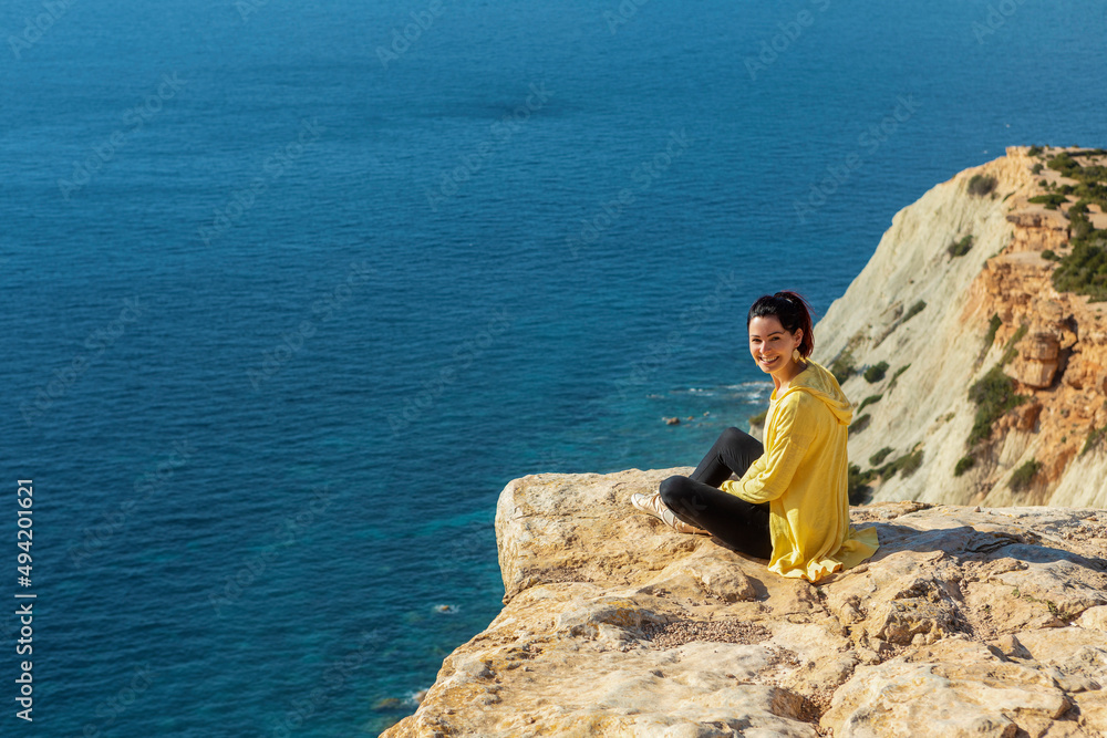 young woman in yellow zip jacket sitting on a cliff turning back to the camera young woman in yellow zip jacket sitting on a cliff turning back to the camera