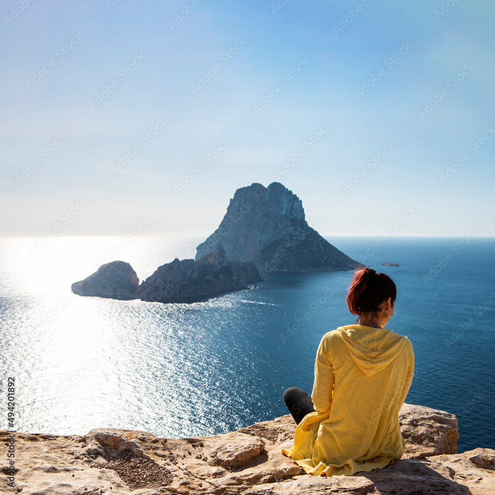young woman in yellow zip jacket sitting on a cliff looking out on the Mediterranean Sea from Ibiza to famous Es Vedra