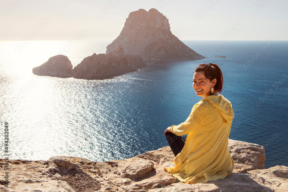 young woman in yellow zip jacket sitting on a cliff turning back to the camera with famous view from Ibiza to Es Vedra in the background