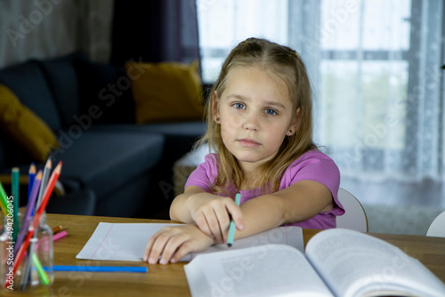 A cute blonde girl thought before completing a school task sitting at a table at home in the living room. In his hand is a pencil on the table notebook. The concept of homeschooling, online education