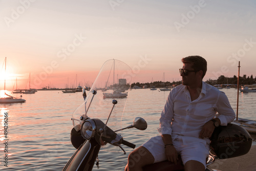 A modern senior businessman in casual clothes sitting on a scooter by the sea and enjoys the sunrise. Selective focus 