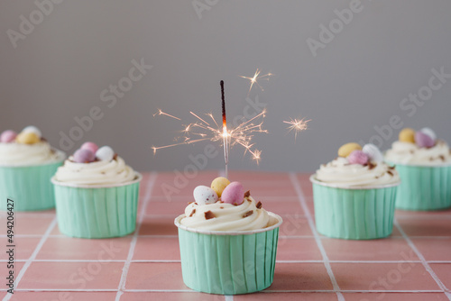 Easter egg cupcakes set against a pale pink tiled work surface and plain background. Cupcake decorated with mini chocolate eggs and cream frosting. Front cupcake has lit sparkler burning.