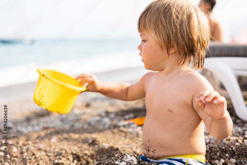 Cute child playing with yellow bucket and toy car on pebble beach. Happy toddler on sea coast. Summer family holiday vacation. Two years old little boy having fun. Summertime. Travel concept Cute child playing with yellow bucket and toy car on pebble beach. Happy toddler on sea coast. Summer family holiday vacation. Two years old little boy having fun. Summertime. Travel concept