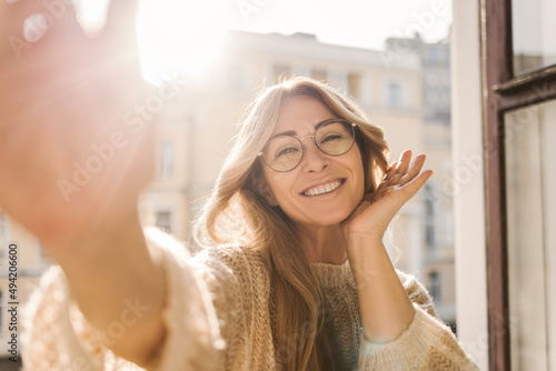 Pretty caucasian adult woman in glasses smiling while looking at camera while standing by open window on sunny day. Blonde in warm sweater spends time alone. Beauty, emotions people concept