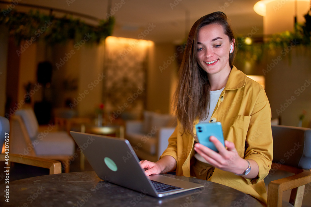 Happy cute joyful modern satisfied smart young casual girl freelancer wearing wireless headphones using phone and laptop for remotely working in a cafe Happy cute joyful modern satisfied smart young casual girl freelancer wearing wireless headphones using phone and laptop for remotely working in a cafe