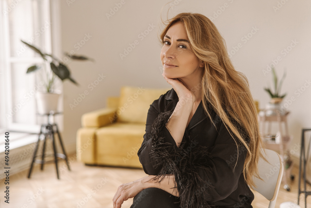 Pretty middle-aged caucasian lady with long blond hair is sitting at home in spacious room. Woman model wears silk black pajamas. Good morning concept