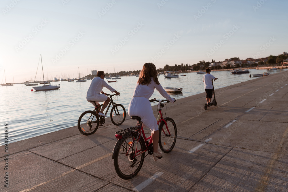 Fototapeta premium Happy family enjoying a beautiful morning by the sea together, parents riding a bike and their son riding an electric scooter. Selective focus 