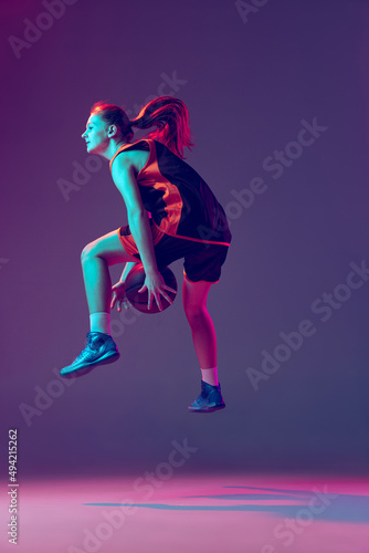 Portrait of young girl, basketball player dribblig ball in jump isolated over gradient pink purple background in neon