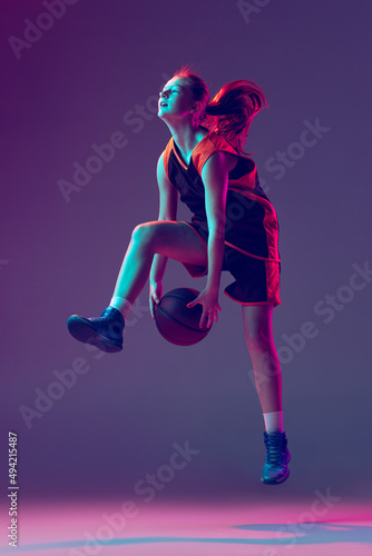 Portrait of teen girl, basketball player dribblig ball in jump isolated over gradient pink purple background in neon © master1305