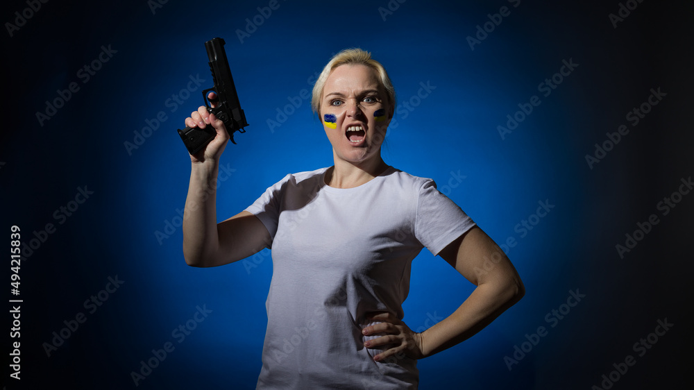 An aggressive woman in a white T-shirt screams with a gun in her hands with the Ukrainian flag on her cheek on a dark background in hard light. Patriot concept. An aggressive woman in a white T-shirt screams with a gun in her hands with the Ukrainian flag on her cheek on a dark background in hard light. Patriot concept.