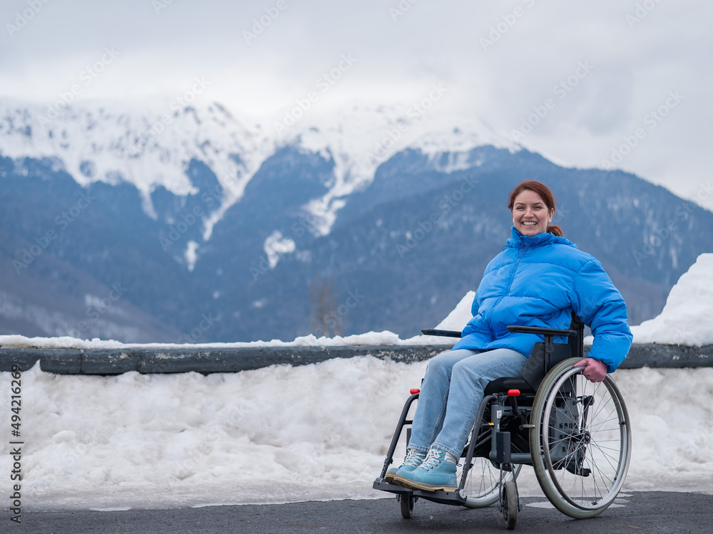 A happy woman dressed in a blue coat sits in a wheelchair on a point view and looking at the snow-capped mountains. 