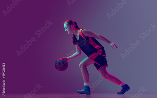 Portrait of young girl, teen, basketball player in motion, training isolated over gradient pink purple background in neon. Side view