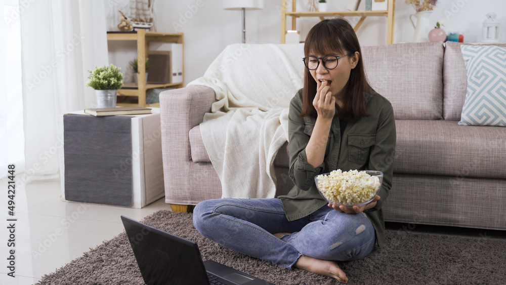 Obraz premium relaxed asian young lady streaming comedy series on notebook and enjoying popcorn in the cozy living room in daytime
