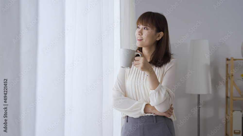 successful smiling asian japanese female entrepreneur sipping tea while looking at view outside office while standing by window. successful smiling asian japanese female entrepreneur sipping tea while looking at view outside office while standing by window.