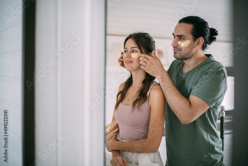 Family spa day at home. Young woman and man with golden hyaluronic patches on their faces in the bedroom of their home.