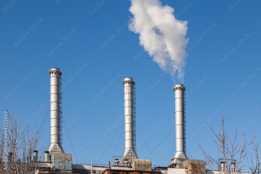 Three factory chimneys against a clear sky. White smoke coming from one of the pipes