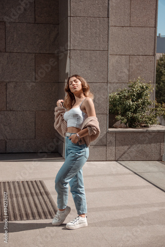 young curly brunette girl in white top and blue jeans is standing fashion near store at sunny day and sunbathing with smile. lifestyle concept, free space © NASTYA PALEHINA