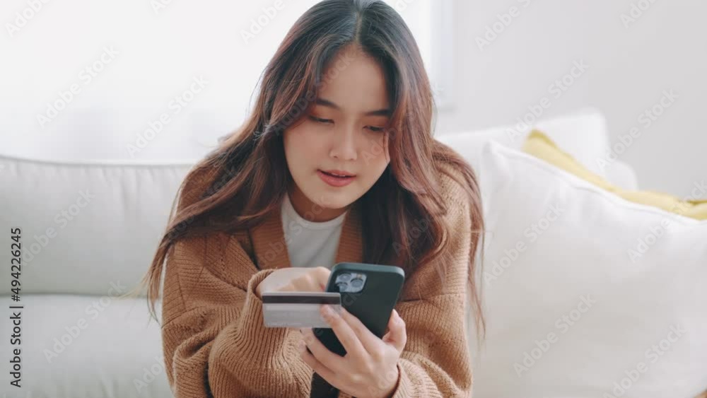 Happy young asian woman holding credit card using instant mobile payments at home. Smiling Female customer shopper making purchase on smartphone receiving cash back concept. E-banking app service. Happy young asian woman holding credit card using instant mobile payments at home. Smiling Female customer shopper making purchase on smartphone receiving cash back concept. E-banking app service.
