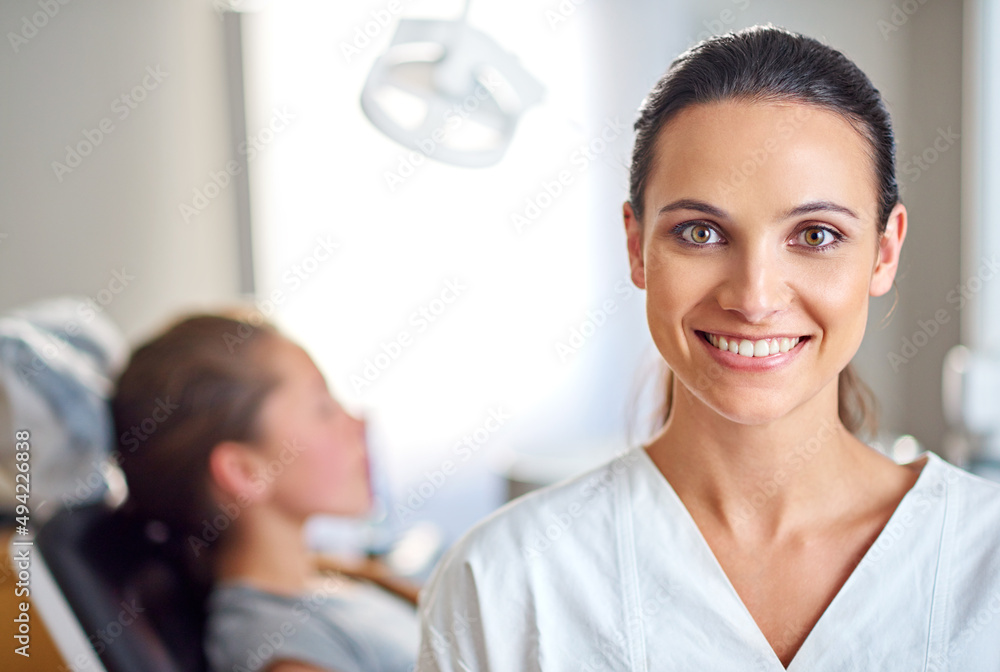 So glad I decided to specialize in dentistry. A female dentist standing in her office with her child patient in the background. So glad I decided to specialize in dentistry. A female dentist standing in her office with her child patient in the background.