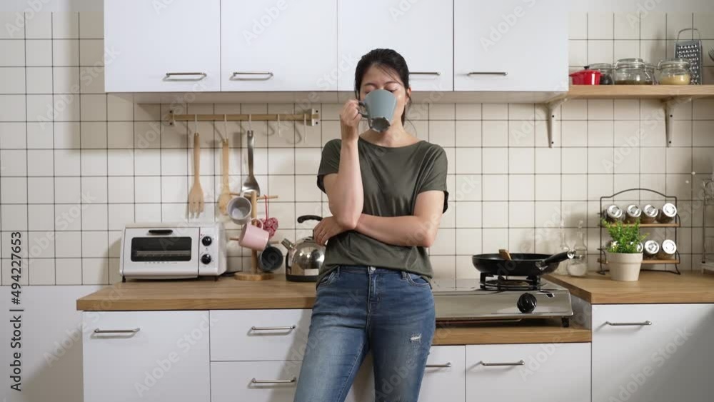 front view of a serious asian woman leaning on kitchen platform and looking into distance with a thoughtful face while drinking coffee in the morning at home. front view of a serious asian woman leaning on kitchen platform and looking into distance with a thoughtful face while drinking coffee in the morning at home.