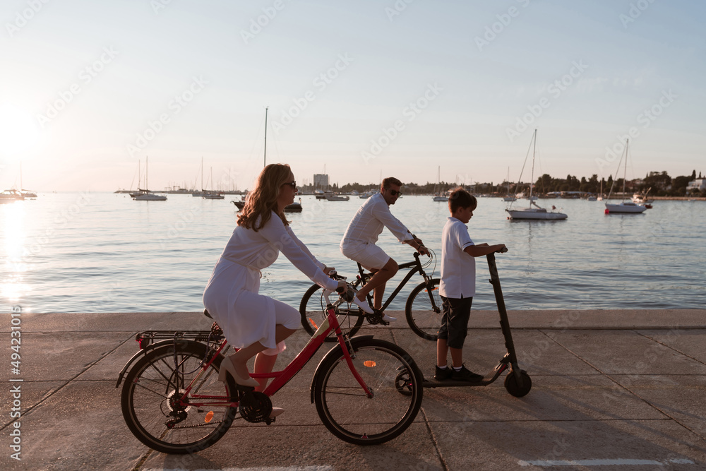 Fototapeta premium Happy family enjoying a beautiful morning by the sea together, parents riding a bike and their son riding an electric scooter. Selective focus 