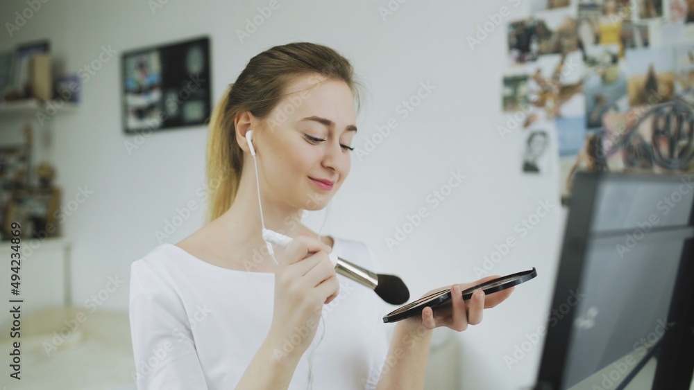 A young woman is sitting at a table in front of a mirror, listening to music with headphones and doing makeup A young woman is sitting at a table in front of a mirror, listening to music with headphones and doing makeup