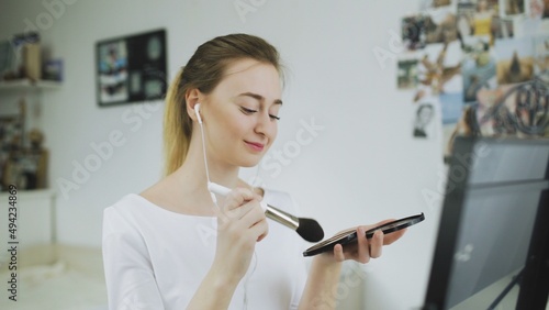 A young woman is sitting at a table in front of a mirror, listening to music with headphones and doing makeup