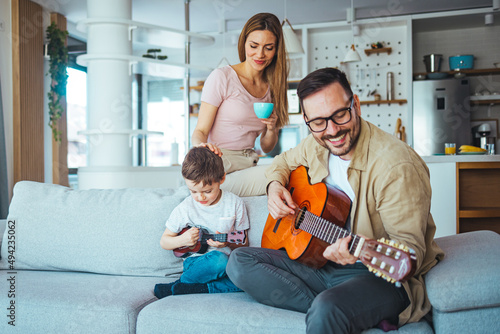 Father teaching his son to play guitar at home. The father teaches his son to play the guitar. He helps him pick up guitar chords. Mom watching. They are in a good mood.