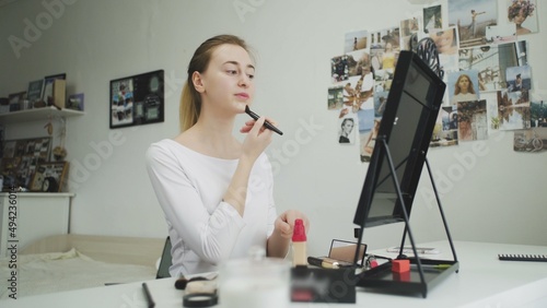 A young woman does her makeup while sitting at a table in front of a mirror