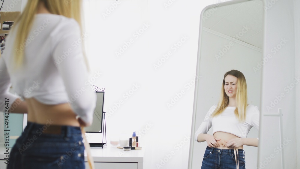 A young woman stands in front of a mirror and measures the circumference of her waist with a tape measure. The girl is complexed because of the weight A young woman stands in front of a mirror and measures the circumference of her waist with a tape measure. The girl is complexed because of the weight