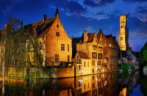 Bruges, Belgium. Evening sunset with blue sky. Water channels of ancient medieval town with view to Belfort van Brugge tower, famous landmark.