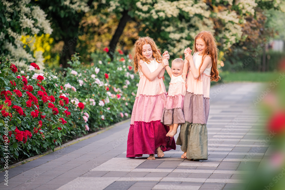 Three red-haired sisters in long linen dresses are merrily walking in the garden with roses on summer evening. Three red-haired sisters in long linen dresses are merrily walking in the garden with roses on summer evening.