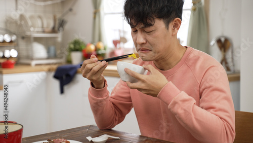 portrait of asian father making expression of disgust while tasting food with chopsticks and bowl at home dining table alone.