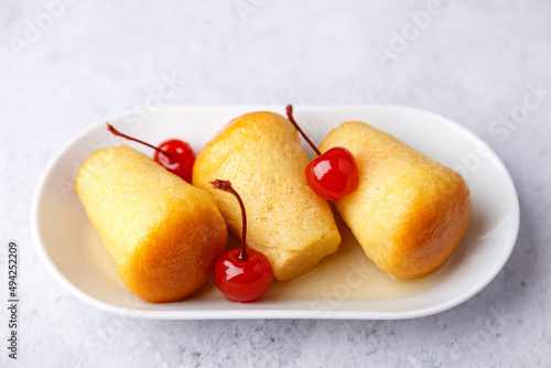 Neapolitan Rum baba (or baba au rum) on a white plate with a cocktail cherry on a gray background. Small yeast cakes soaked in rum syrup. Traditional Italian pastry. Close-up, selective focus.