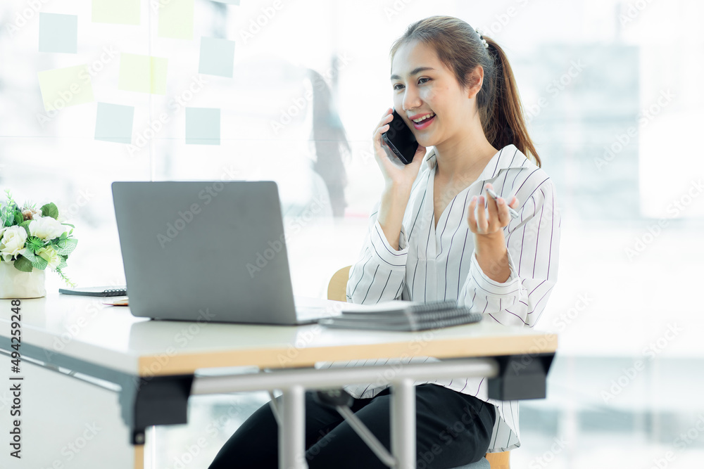 Cheerful young Asian woman smiling and looking away while having smartphone conversation in office Cheerful young Asian woman smiling and looking away while having smartphone conversation in office
